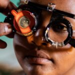 Close-up of the face of Nigest, a young Ethiopian girl as she undergos a vision test. She is wearing a trial frame fitted with test lenses, including a prominent red lens on the left side. A hand adjusts the frame gently. Nigest's expression is calm and focused.