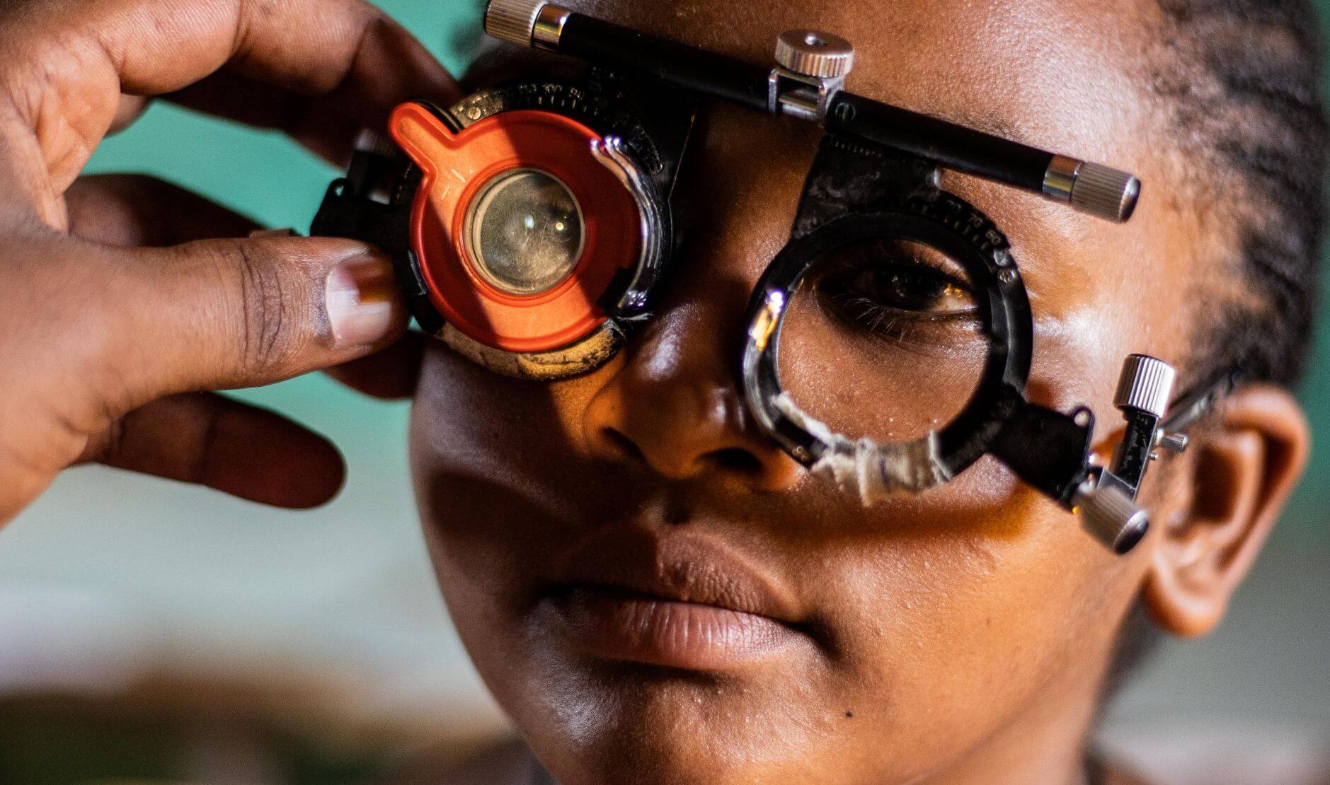 Close-up of the face of Nigest, a young Ethiopian girl as she undergos a vision test. She is wearing a trial frame fitted with test lenses, including a prominent red lens on the left side. A hand adjusts the frame gently. Nigest's expression is calm and focused.