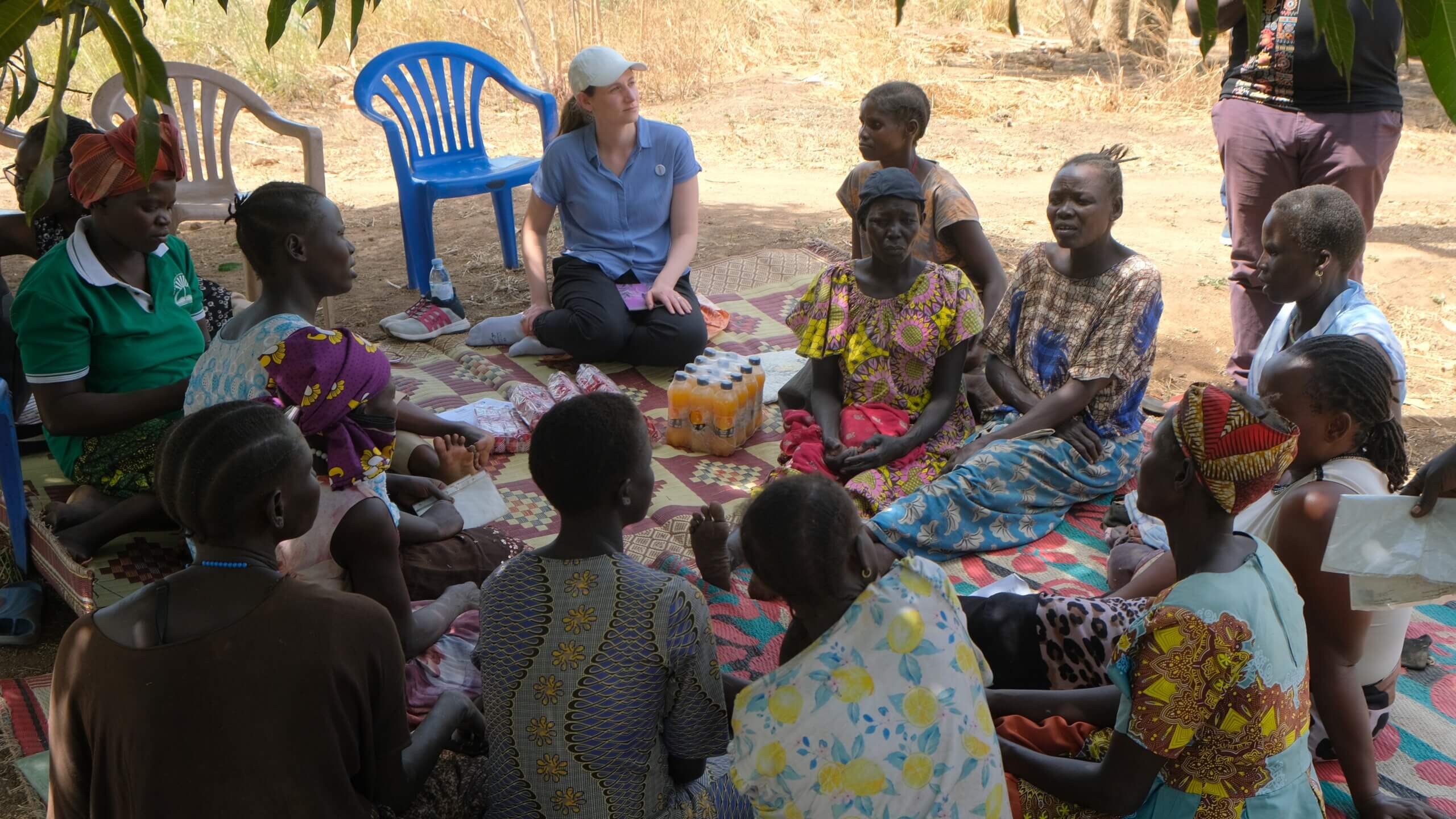 A group of people sit on mats outdoors in a circle under the shade of trees. They are engaged in a discussion with a representative from Light for the World. The scene is part of the Resilience Project in Palabek, Lamwo District, Uganda.