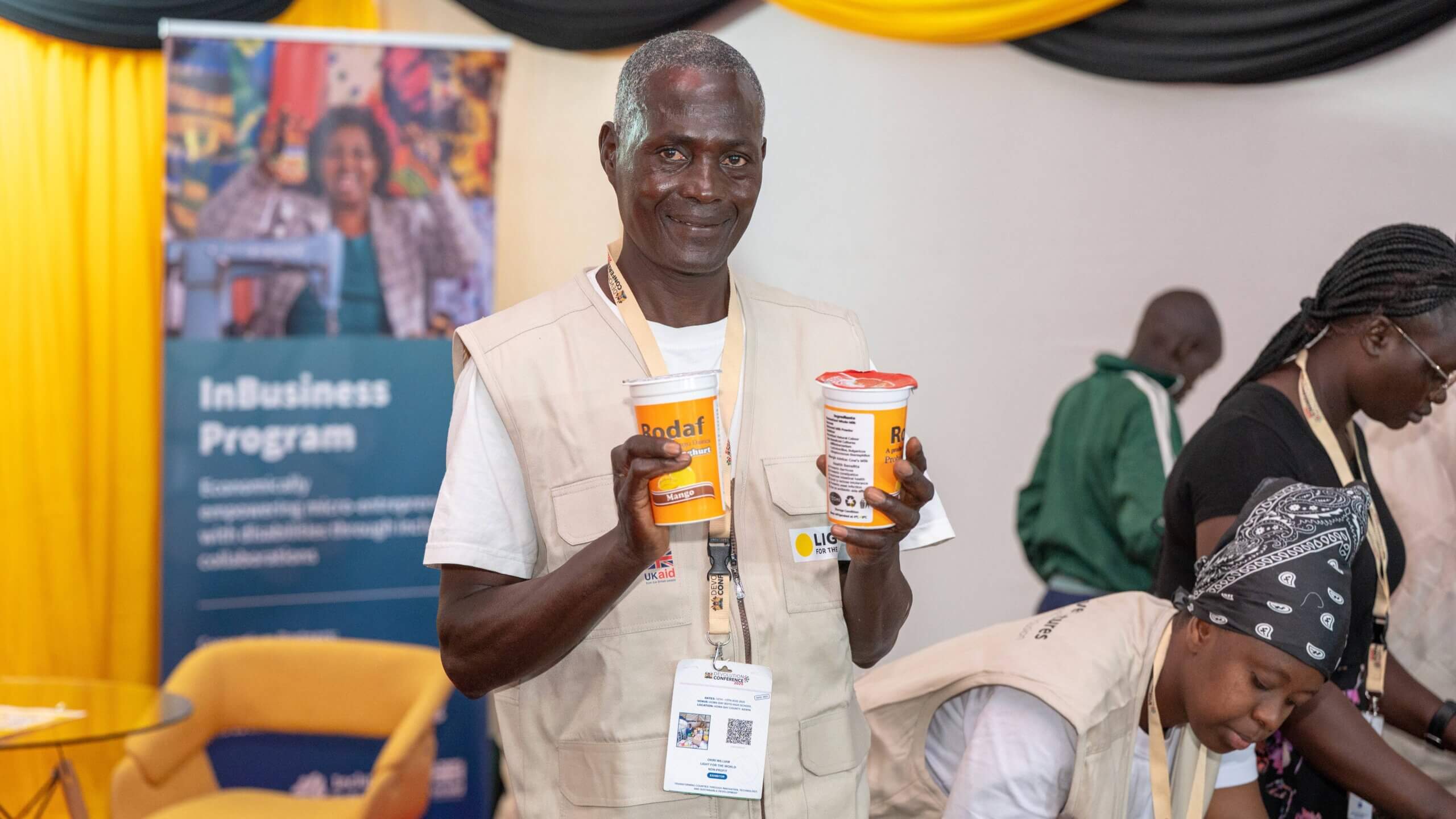 William Okiri, a micro-entrepreneur from Homabay County, holds two containers of yoghurt in front of a banner labeled "InBusiness Program" at the Devolution Conference 2025. The event focused on advancing disability inclusion in employment and entrepreneurship.