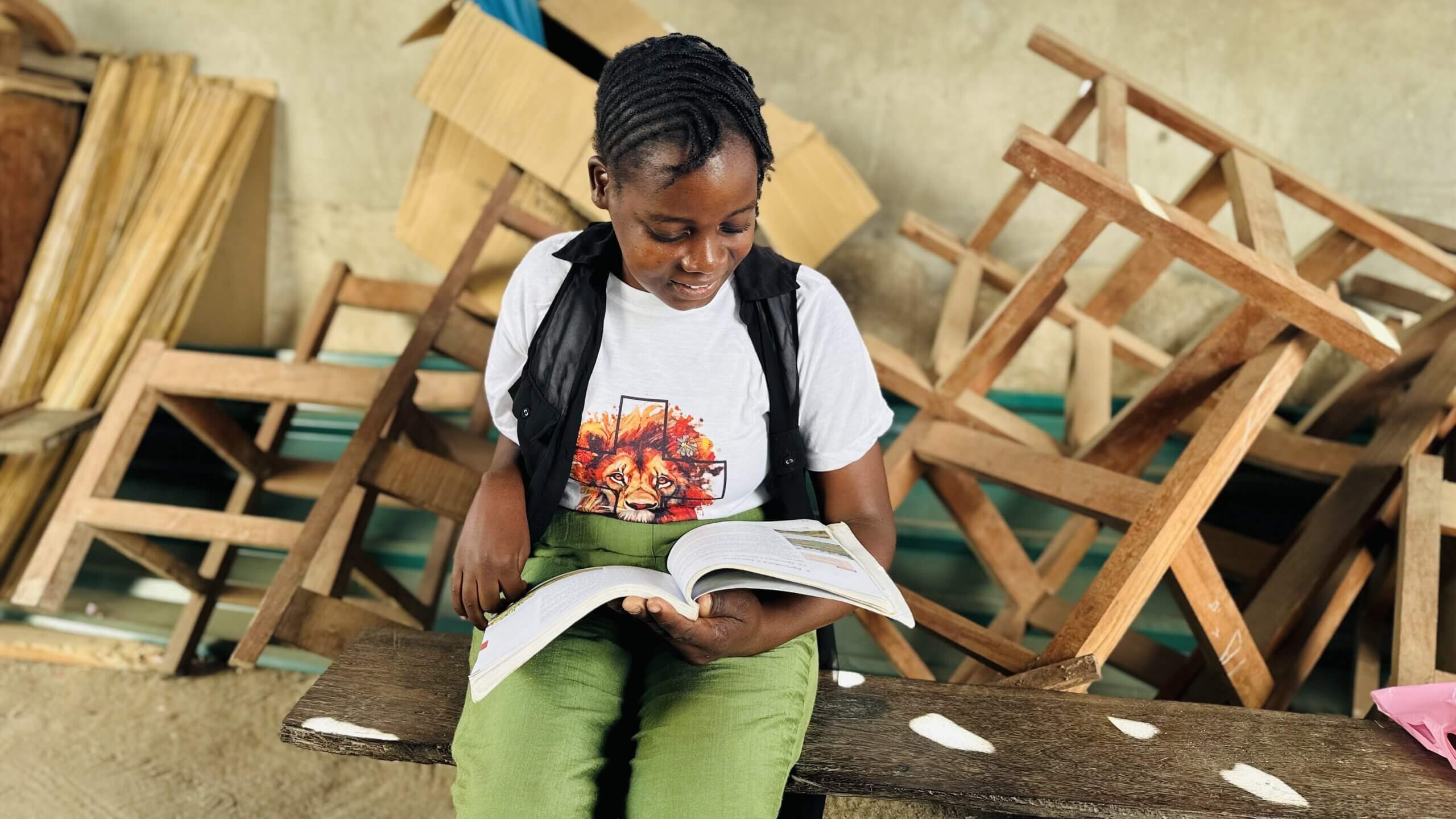 A woman sits on a bench holding an open book. She wears a white t-shirt with a lion design and green pants. Her face is blurred for privacy. Behind her are stacked wooden chairs and furniture against a wall.