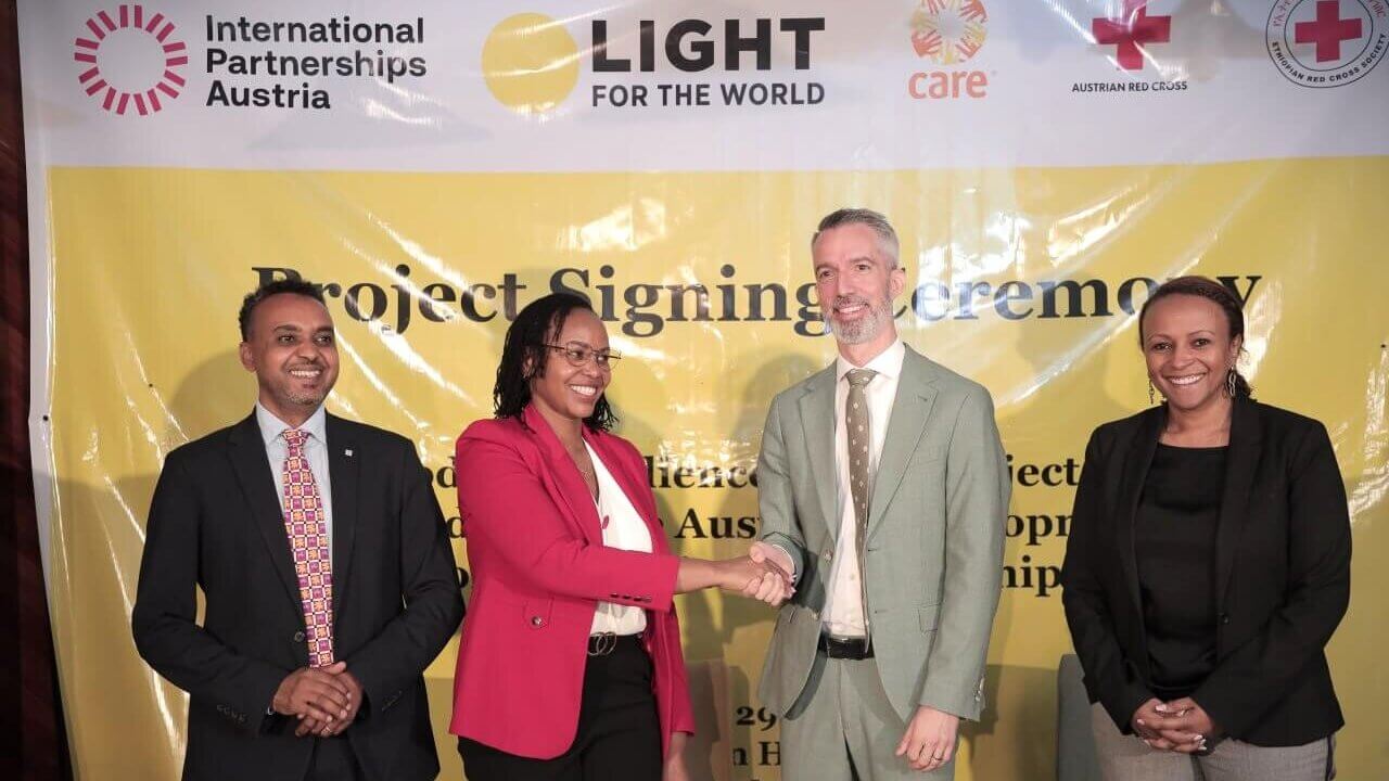 A group of four individuals stand in front of a yellow banner with logos of International Partnerships Austria, Light for the World, CARE, and Austrian Red Cross. Two individuals in the center are shaking hands. The event is identified as a project signing ceremony.