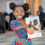Alice Mbere, a Trainee Disability Inclusion Facilitator in Kenya, smiles while holding up a Disability Inclusion Advisory Service Resource Book. She is wearing a bright dress of red and blue.