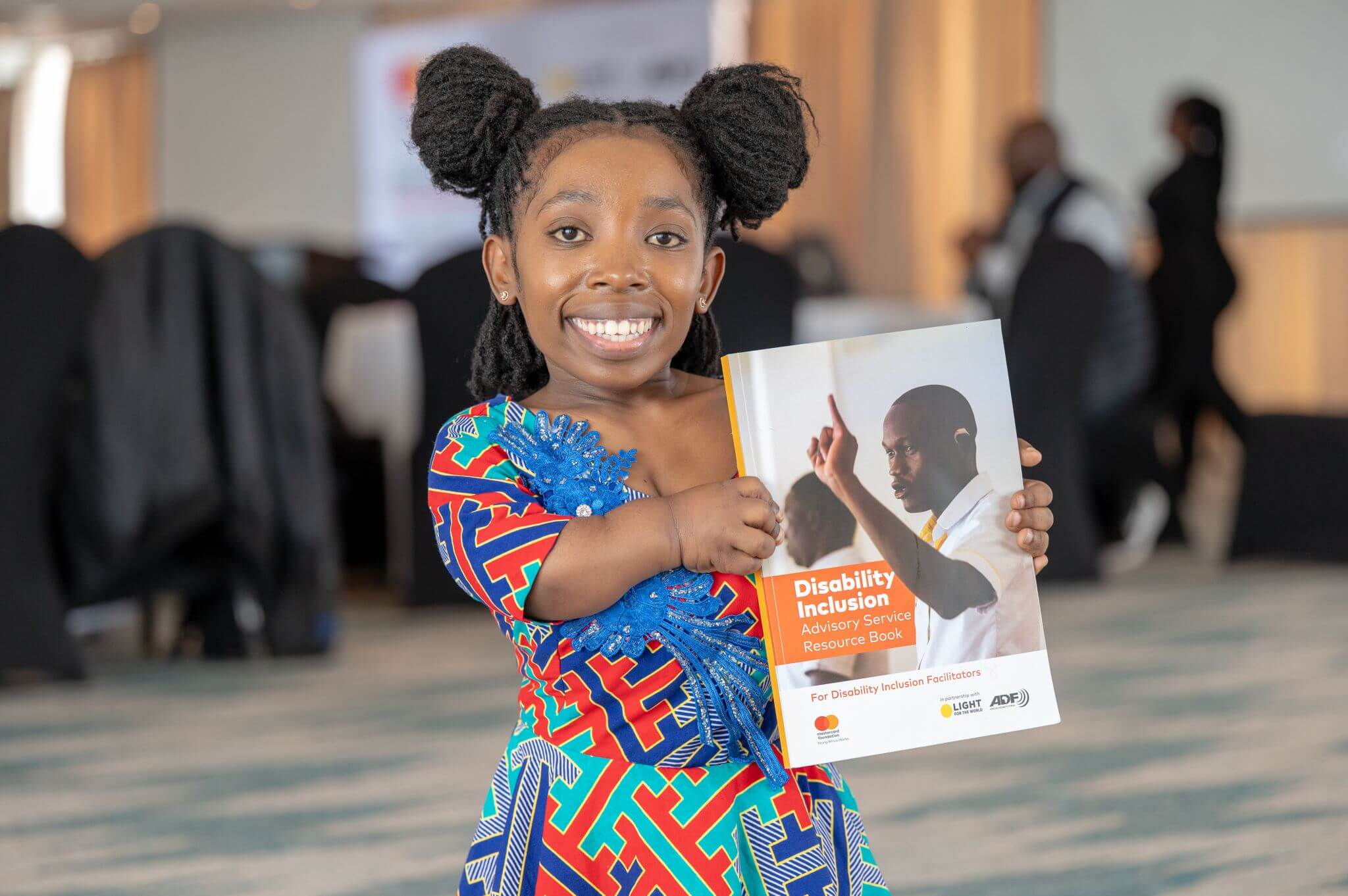 Alice Mbere, a Trainee Disability Inclusion Facilitator in Kenya, smiles while holding up a Disability Inclusion Advisory Service Resource Book. She is wearing a bright dress of red and blue.