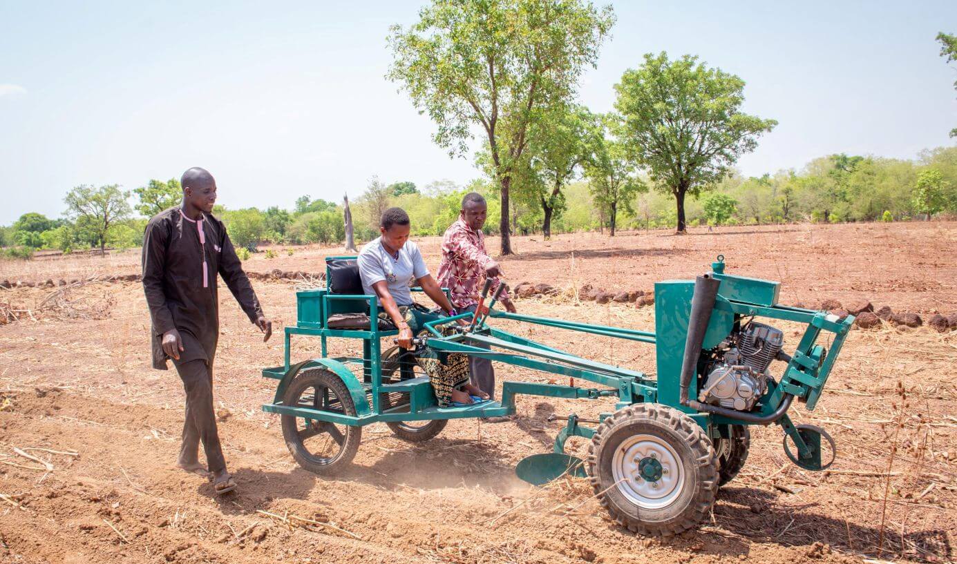 Caroline Somé, a farmer in Burkina Faso, operates a modified cultivator in a field during an AgriLabs workshop on disability-inclusive farm tools. Two men walk alongside the machine with one advising Caroline on how it works.
