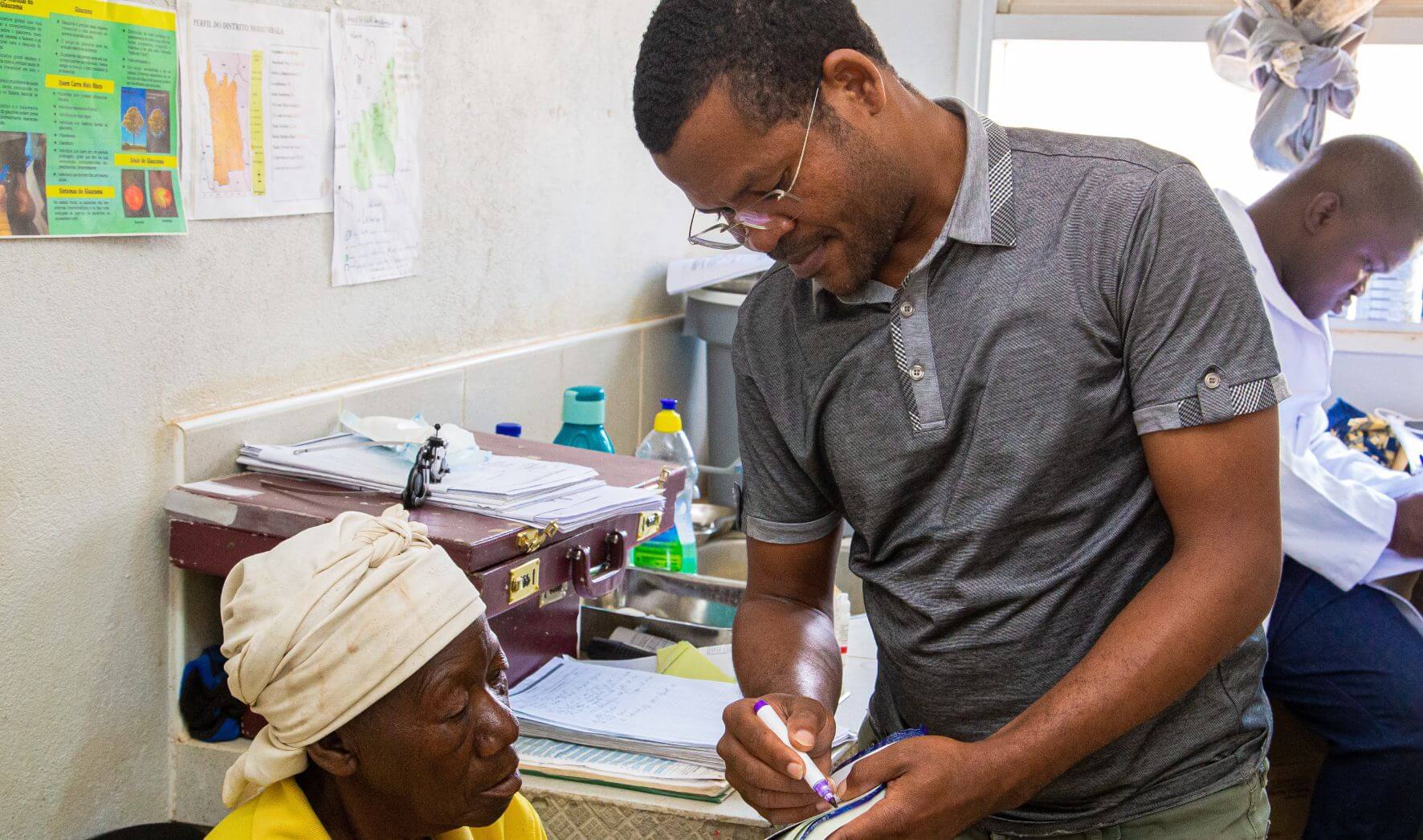 Veronica Mussuba Mambucha, a 69-year-old farmer from Mozambique, in a clinic before receiving a cataract surgery. A staff member is showing her a document and marking it with a pen. In the background, a doctor is present. 