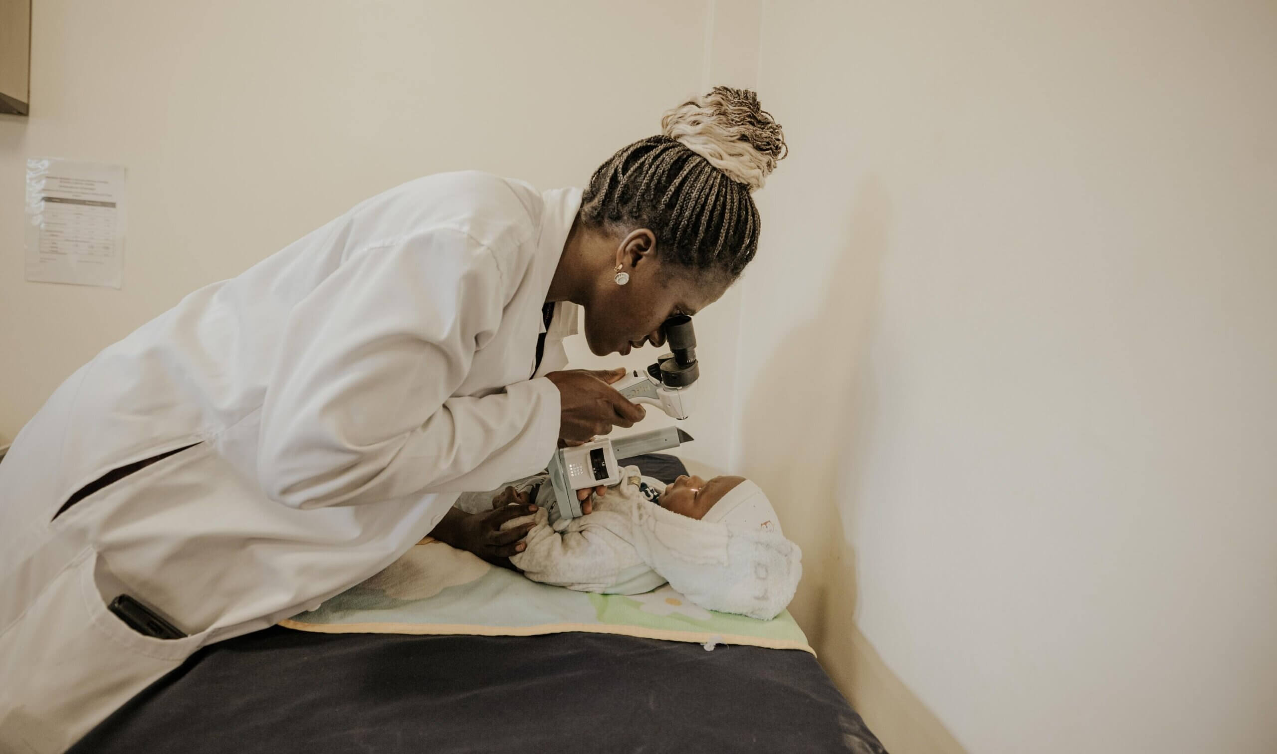 Dr Lusobya, wearing a white doctor's coat, examins the eye of a baby boy with a specialist piece of equipment. They are in her office and the boy lies on an examining table.
