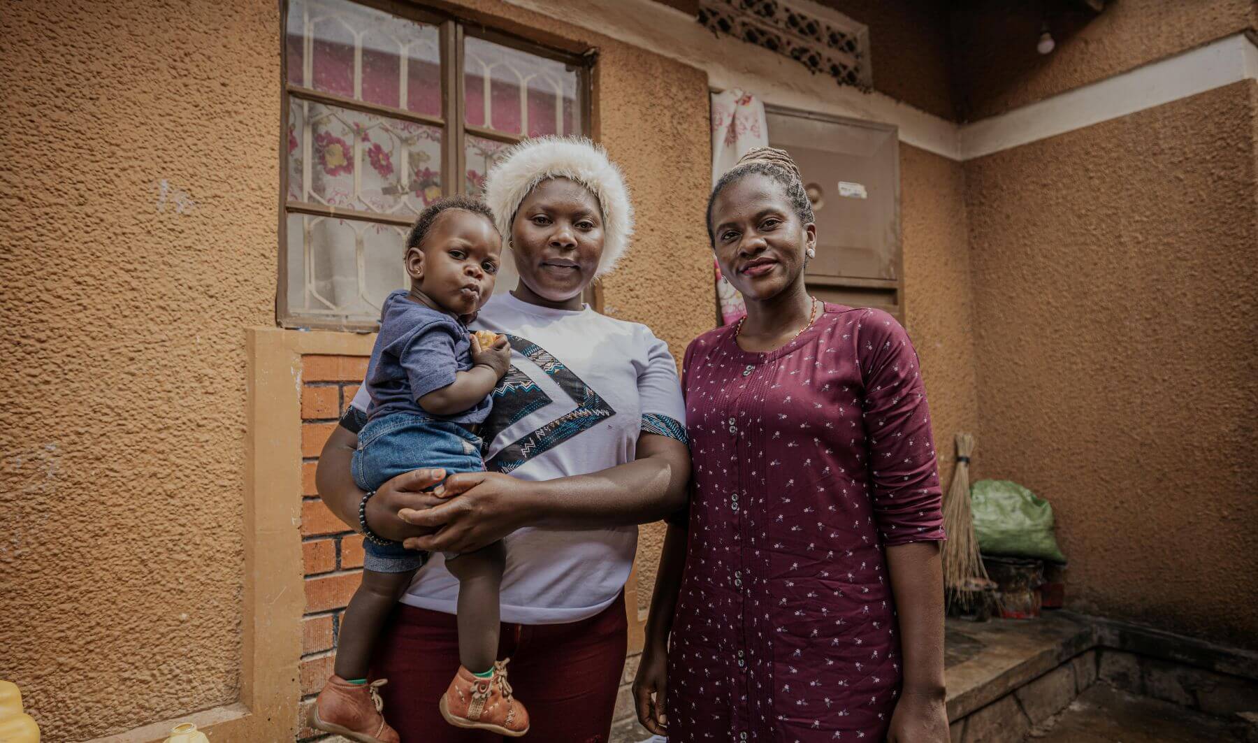 Dr Lusobya, wearing a red dress, stands next to Faziirah Kabahinda, who is holding her one-year-old daughter, Elysh Ahumuza. They are standing in front of a small brick building and all three are looking at the camera with neutral expressions.