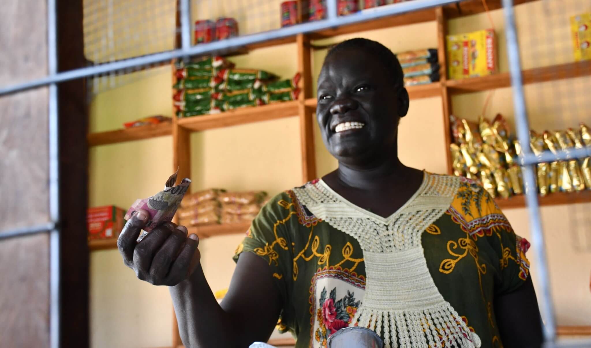 Akuch Deng stands inside her shop holding currency notes, smiling. Shelves behind her are filled with packaged goods. She received business training through the We Are Able! programme and now runs the store with her daughter, promoting disability-inclusive entrepreneurship.