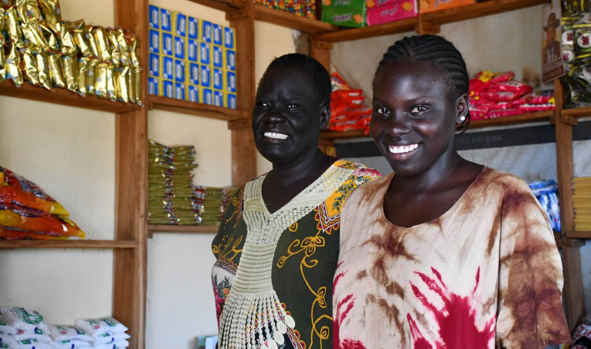 Akuch Deng and her daughter Nyabeny stand inside a small general store in Wau, South Sudan. Shelves behind them are stocked with household goods like rice and soap. They are smiling, reflecting their success in running the business together after receiving training through the We Are Able! programme.