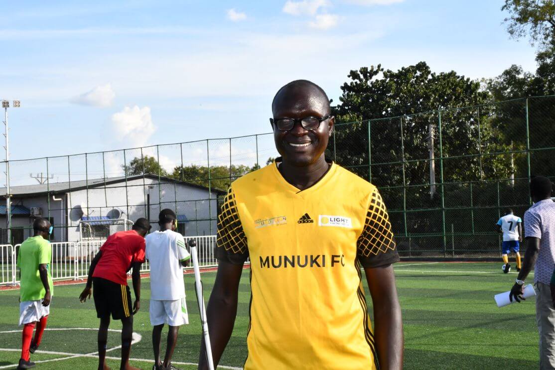 A man wearing a yellow “MUNUKI FC” jersey smiles at the camera. He is standing outdoors and is involved in blind football leadership and legal inclusion advocacy.