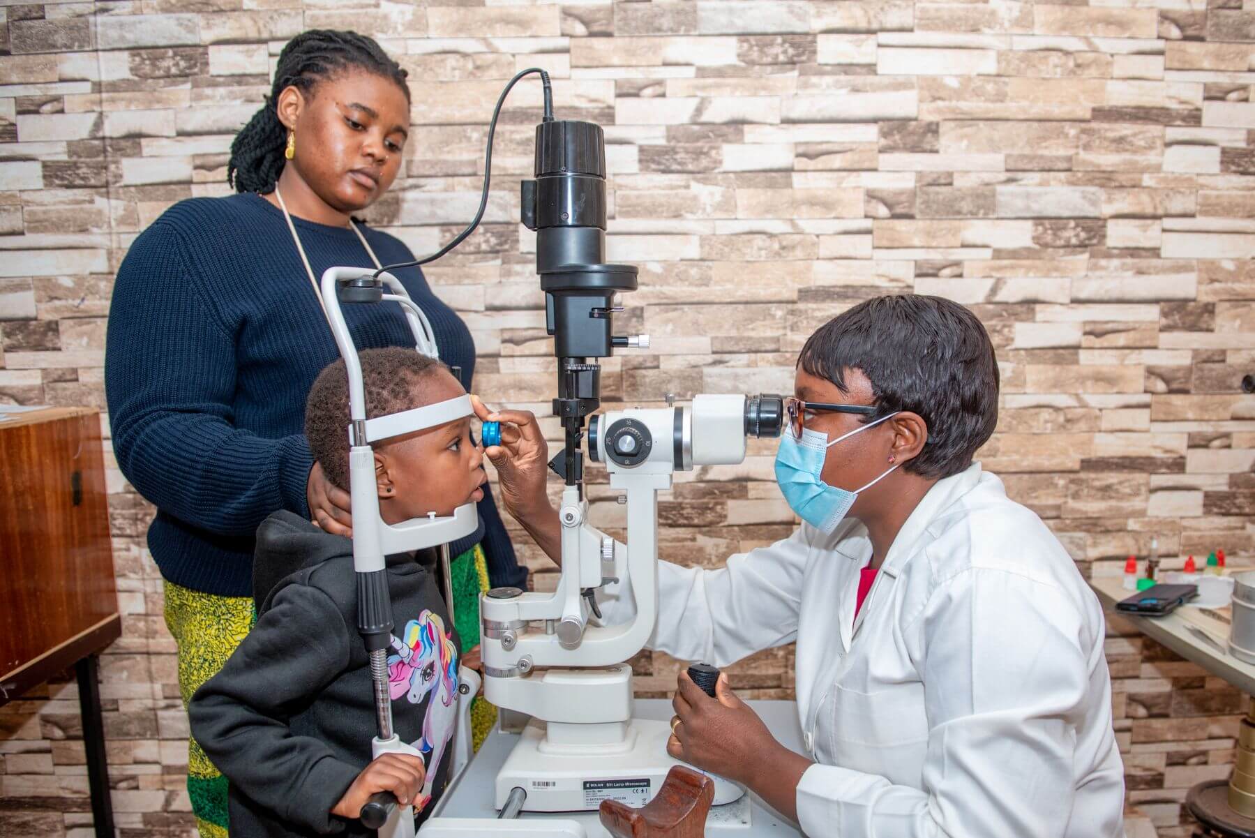 Dr Yaméogo testing the eyes of a four-year-old patient using a machine. The patient's mother is holding her head gently.