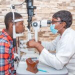 Dr Claudette Yaméogo, in a white doctor's coat and wearing a face mask, tests the eyes of a teenage patient. THey sit either side of the machine and are both looking in the lenses.