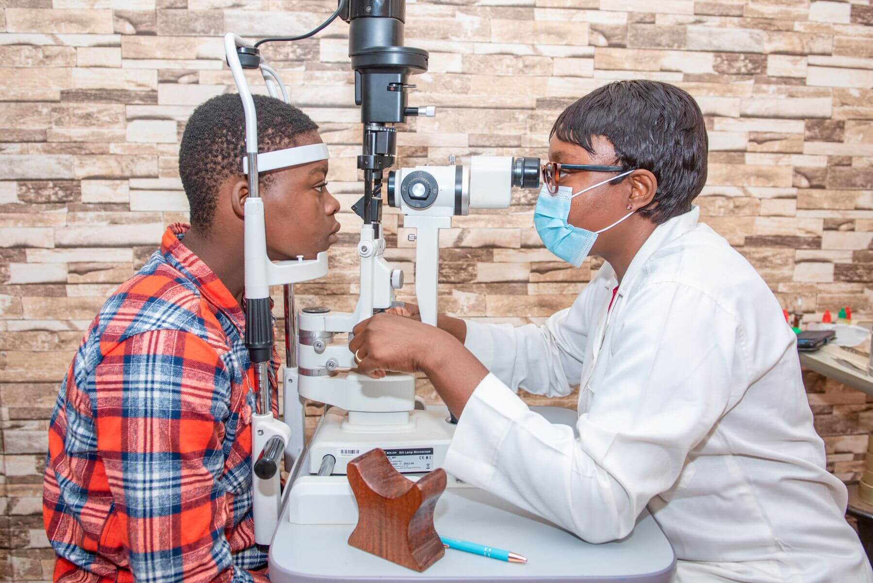 Dr Claudette Yaméogo, in a white doctor's coat and wearing a face mask, tests the eyes of a teenage patient. THey sit either side of the machine and are both looking in the lenses.