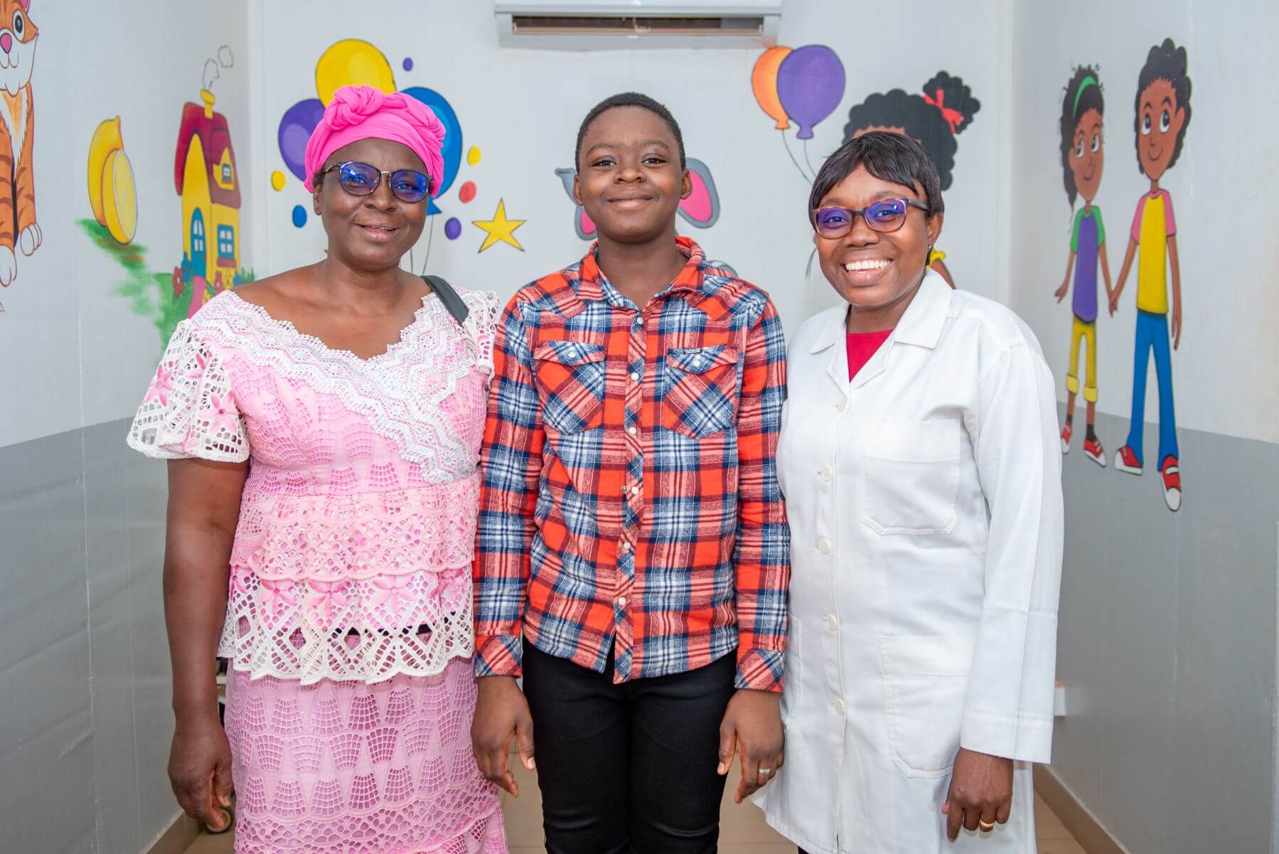 Dr Yaméogo stands with her patient and their mother. All three are smiling. They are in a room where children are treated, with colourful cartoons on the wall in the background.