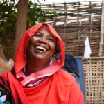 Fatuma Mahamoud sits outdoors, smiling broadly. The background includes greenery and woven wooden panels. She is a mother of five who expanded her business after joining a Village Savings and Loan Association through the We Are Able! programme.