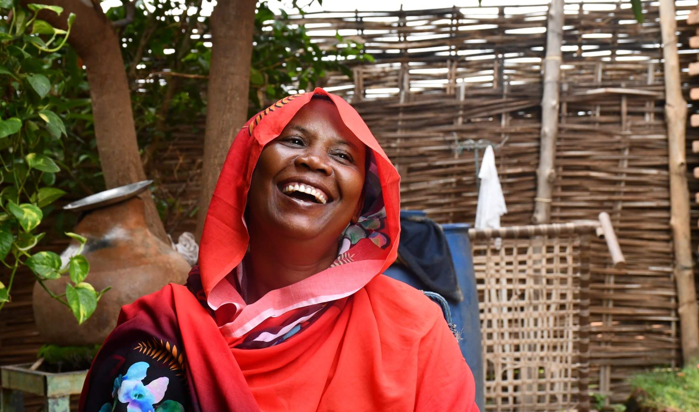 Fatuma Mahamoud sits outdoors, smiling broadly. The background includes greenery and woven wooden panels. She is a mother of five who expanded her business after joining a Village Savings and Loan Association through the We Are Able! programme.
