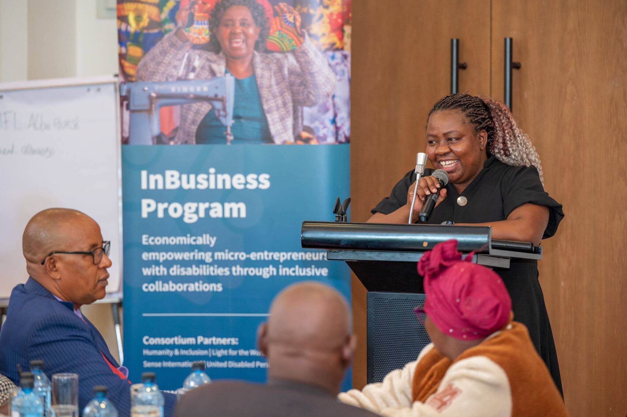 A woman stands at a podium, holding a microphone. She is wearing a black dress and has pink and blonde curls tied back in a pony tail. She has a big smile on her face. Three audience members side in front and there is an "InBusiness Program" sign to her left. It seems like a conference room setting.
