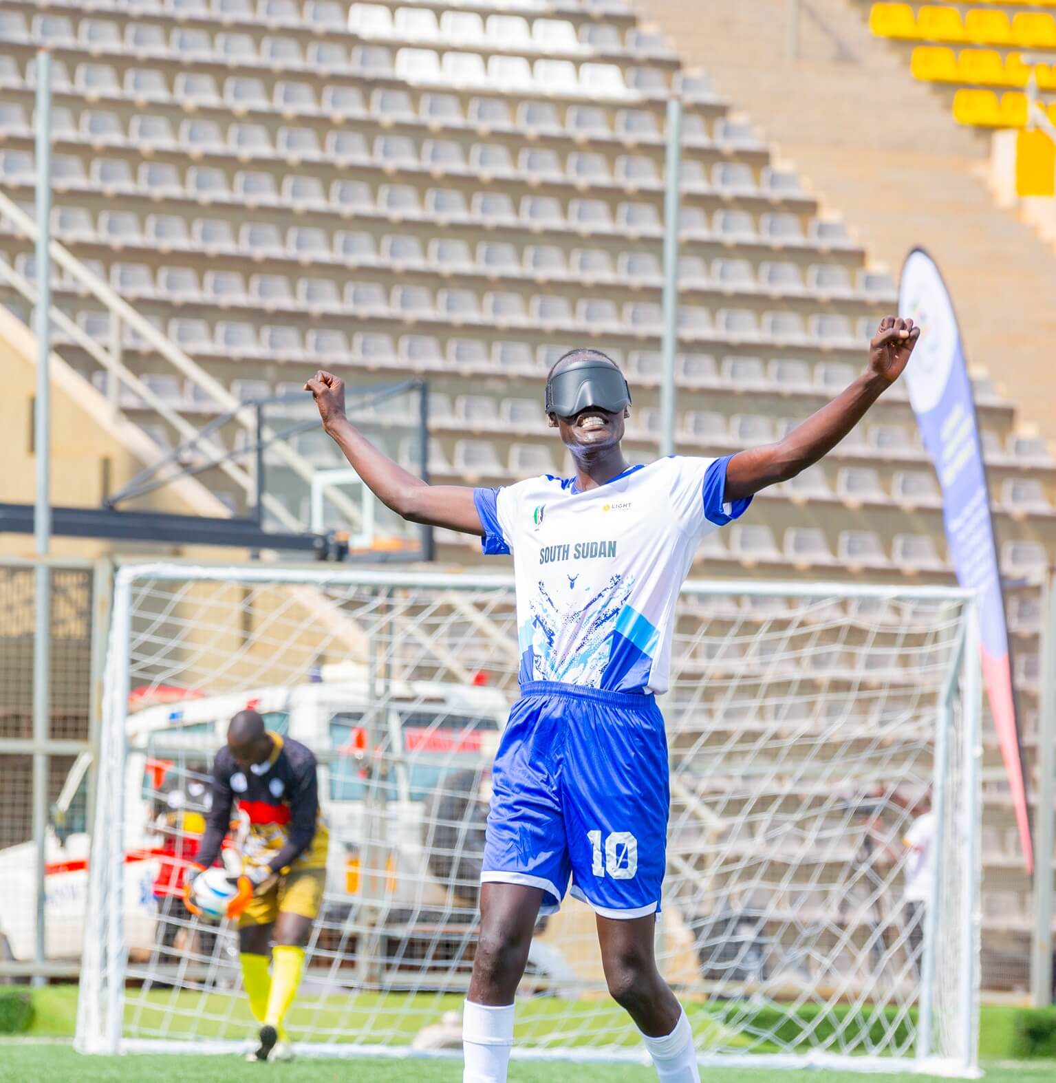 South Sudan captain Martin Ladu Paul in a white and blue kit stands with arms raised in celebration after scoring a goal. Behind, the Uganda goalkeeper in yellow and black prepares to pick up the ball.