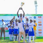South Sudan players in white and blue kits lift the championship trophy high on the podium. Medals hang around their necks as confetti sprays in the air.