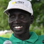 James Mach Nai wearing a white cap with the words “Light for the World” speaks into a microphone outdoors. Trees are visible in the background. He is a disability rights advocate and leader of a savings group, supported by the We Are Able! programme.