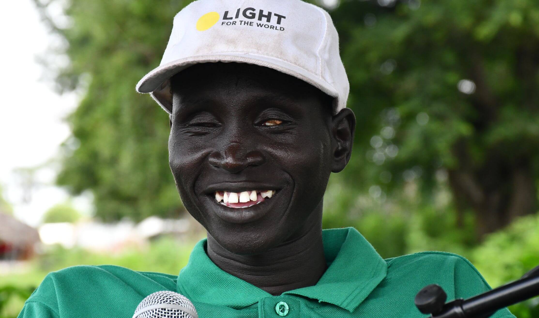 James Mach Nai wearing a white cap with the words “Light for the World” speaks into a microphone outdoors. Trees are visible in the background. He is a disability rights advocate and leader of a savings group, supported by the We Are Able! programme.