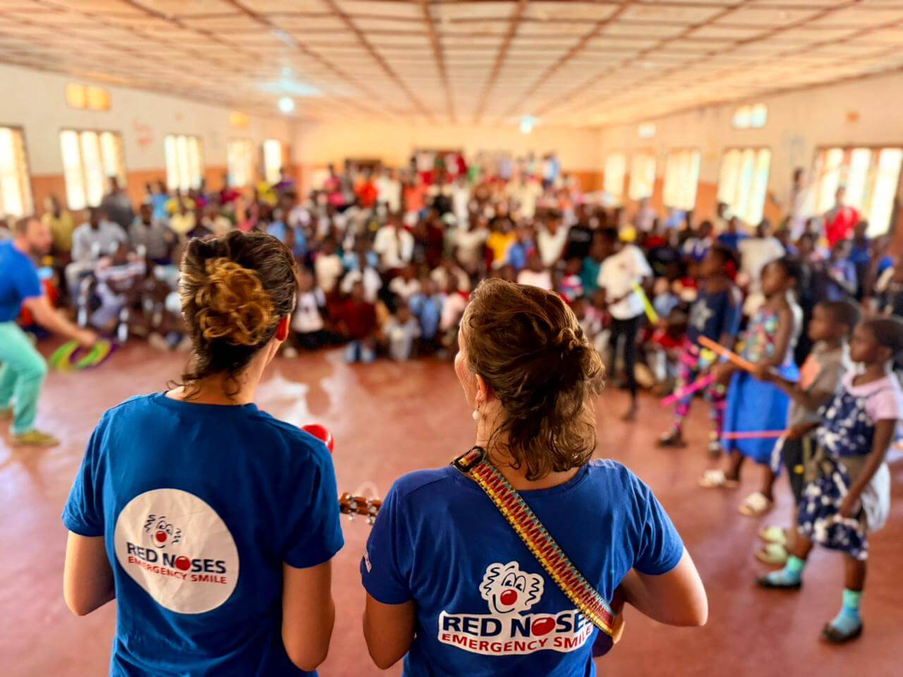 Two women stand with their backs to the camera, wearing a "Red Noses Emergency Smile" blue t-shirt. One is holding a ukulele. A crowd of children and adults are watching, blurred in the background. Some children are standing and taking part in a game. The setting looks like a school hall or classroom.