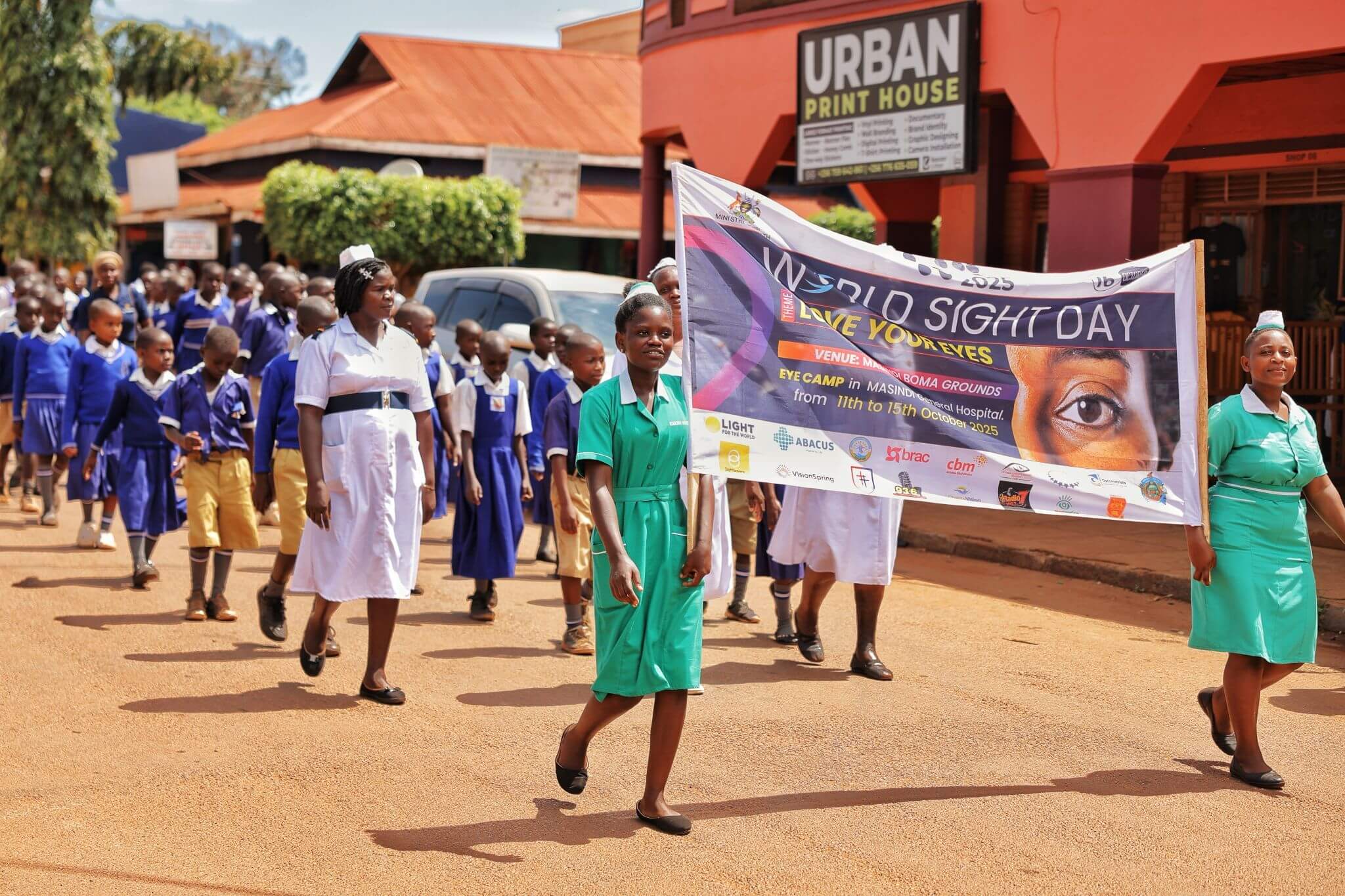 A group of people are walking a parade, Two women in green medical uniforms stand at the front holding a sign that reads "World Sight Day. Love Your Eyes." There are lots of logos including Light for the World. A few more medical staff stand behind in white uniforms. Behind are at least 50 children in blue school uniforms, but the parade ends off of the picture. It is sunny, there is greenery around and rusty orange buildings in the background.