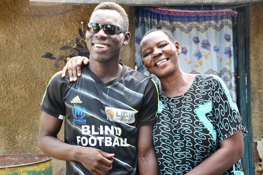 Yona and his mother, Leya Tabu, at their home in Juba. Both are smiling at the camera. Yona is wearing sunglasses and a football shirt that says "Blind Football"