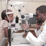 A healthcare professional in a white coat conducting a postoperative eye check-up on a child using an ophthalmic device in a clinical setting.
