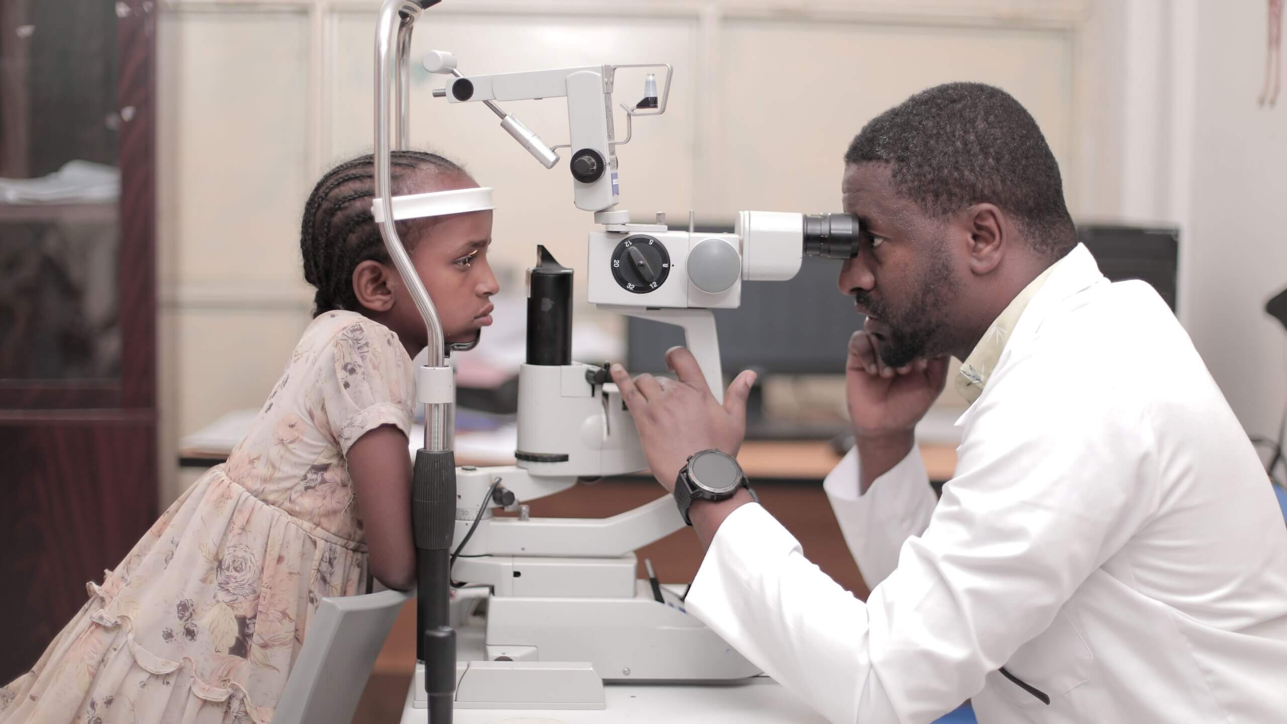 A healthcare professional in a white coat conducting a postoperative eye check-up on a child using an ophthalmic device in a clinical setting.