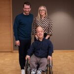 Three people are indoors against a plain wall with a wooden door on the left. Andreas Onea and Marion Lieser are standing side by side, while Tom Shakespeare is seated in a wheelchair in front of them. The floor is light-colored wood, and the setting appears to be a spacious room with neutral tones.