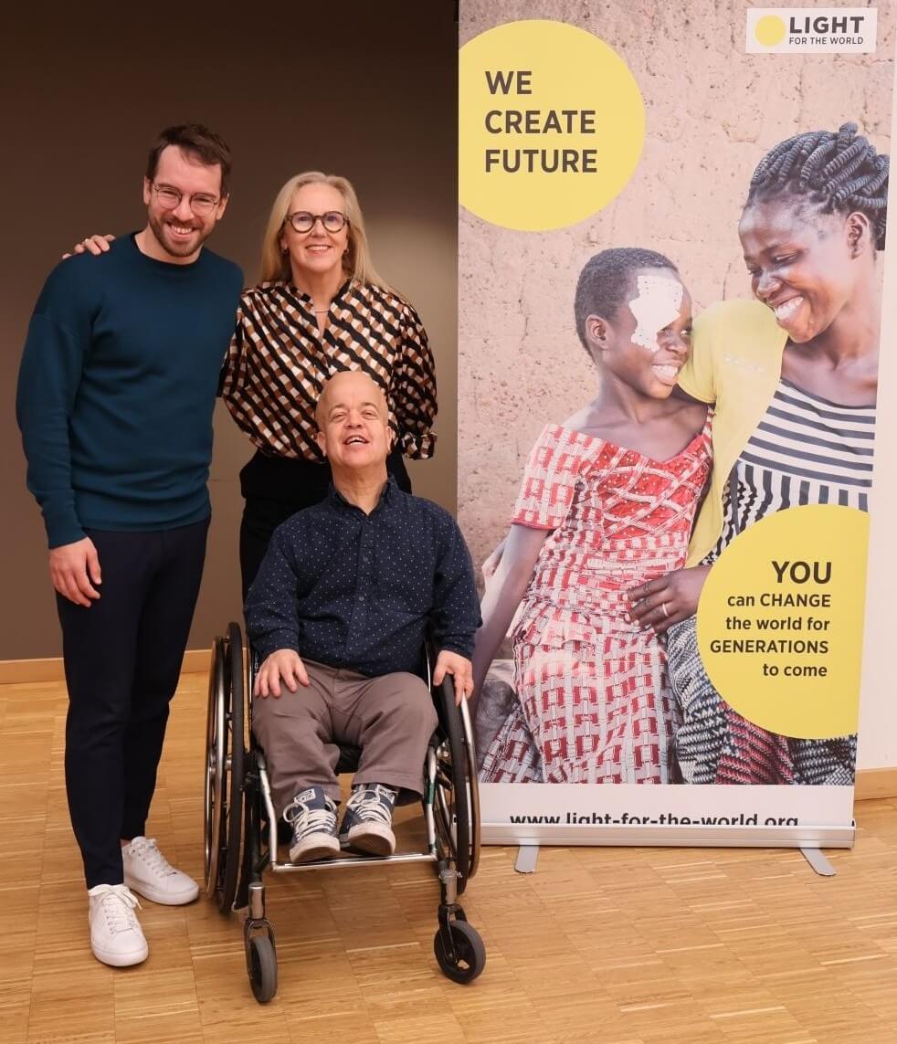 Three people are indoors against a plain wall. A banner from Light for the World is next to them. It has a photo of two smiling women and the words "We create future". Andreas Onea and Marion Lieser are standing side by side, while Tom Shakespeare is seated in a wheelchair in front of Marion. The floor is light-colored wood, and the setting appears to be a spacious room with neutral tones.