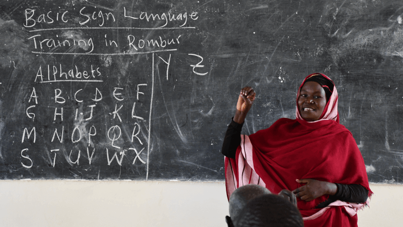 Hanadi, a trainee teacher, practises signs during a class. She is in front of a blackboard and wearing a red dress.