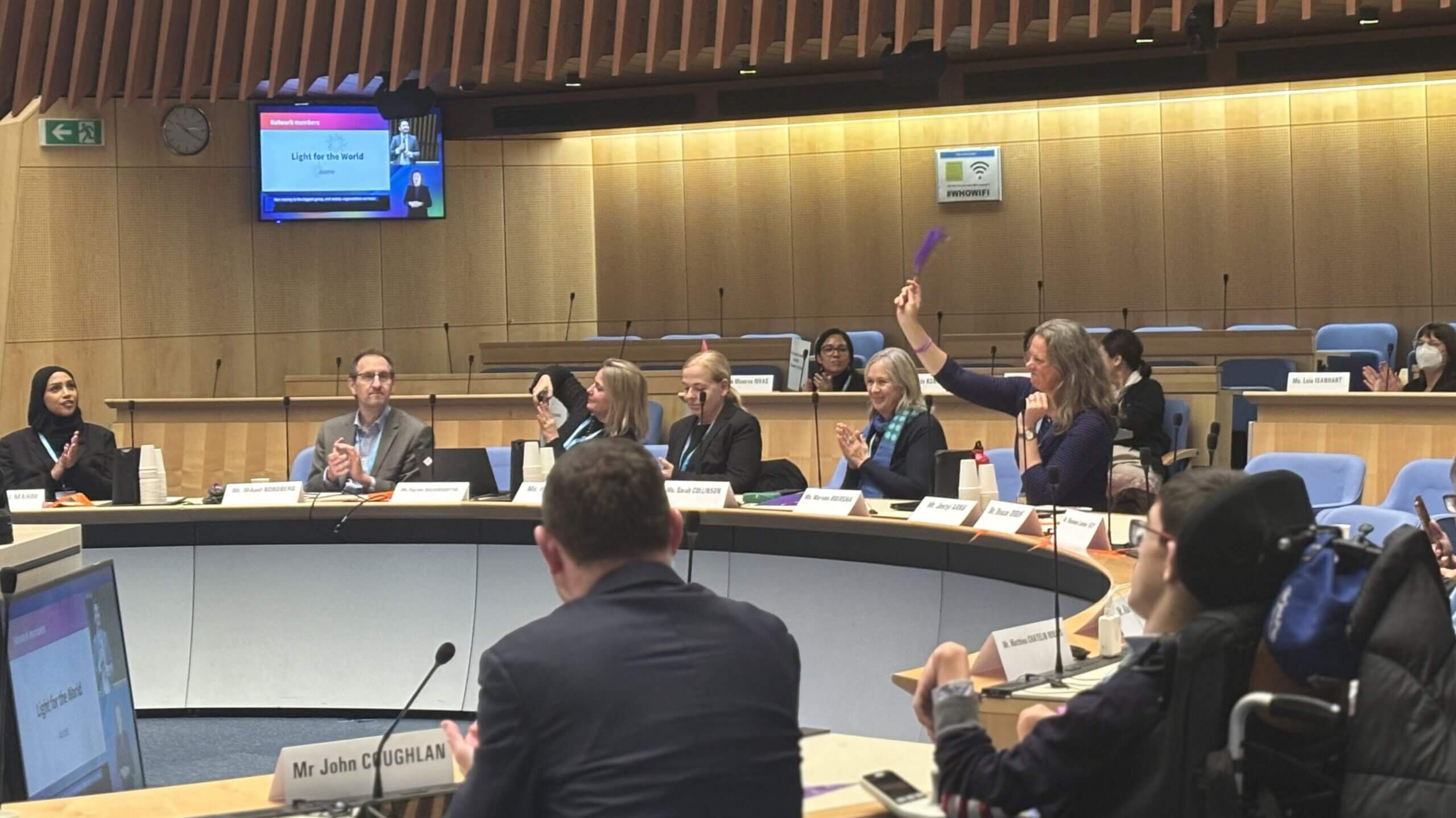 A formal meeting in a modern conference room with people seated around a curved table, nameplates in front of them, and a large screen displaying the text ‘Lights for the World.’