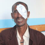 Person sitting on a wooden bench against a light blue and white wall, wearing a brown jacket over a beige shirt, with a white gauze bandage over one eye and smiling at the camera.