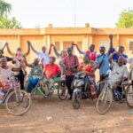 A group of project participants from Burkina Faso and project staff join hands and smile. Some are sat in tricycles.