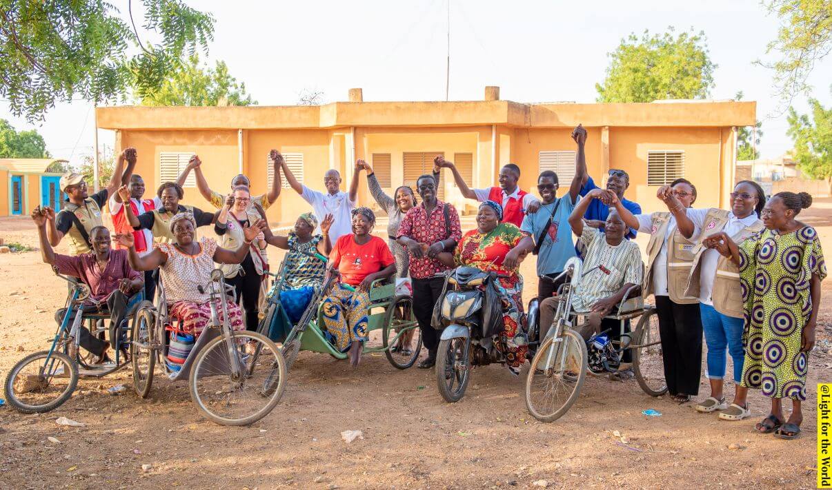 A group of project participants from Burkina Faso and project staff join hands and smile. Some are sat in tricycles. 