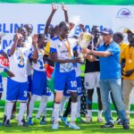 South Sudan players in white and blue kits celebrate as captain Martin Ladu Paul is presented with the championship trophy. Medals hang around their necks, players hold their hands aloft in celebration and everyone is smiling.
