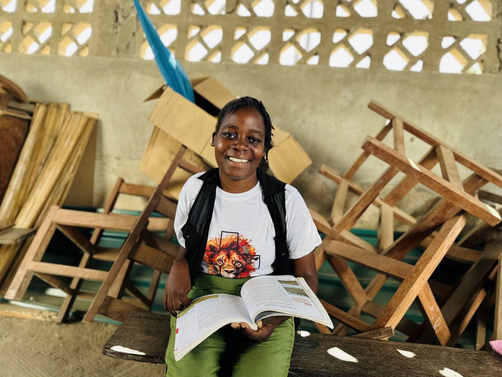 Wilsa Chitato Manuel, a 20-year-old woman from Mozambique, sits in a classroom and smiles at the camera. An open textbook is in her lap. She sits in a classroom and chairs cover the space in the background.