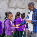 A class of school children receive medication during a MDA campaign in Tigray, Ethiopia. A girl wearing a purple shirt holds out her hand to receive the tablets from a man in a white shirt and blue checked top. There are other children in the background wearing purple school uniforms, with green skirts.