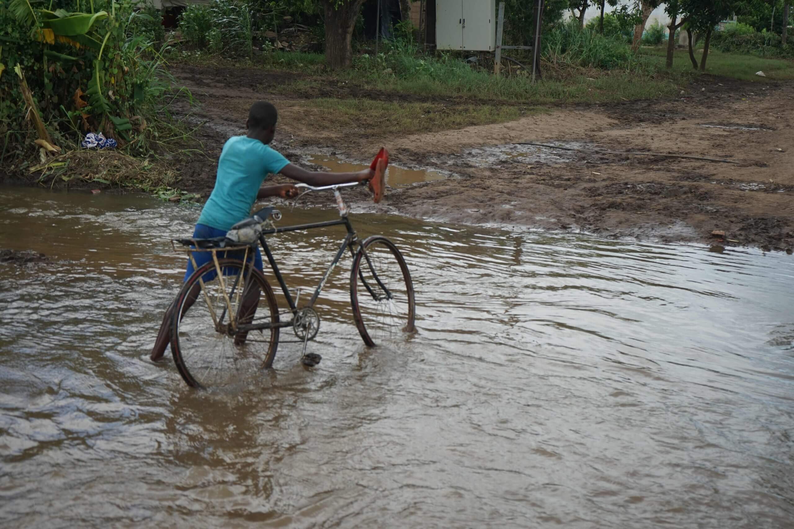 A boy wearing blue clothes pushes a bicycle over a flooded road in Buzi, Mozambique.