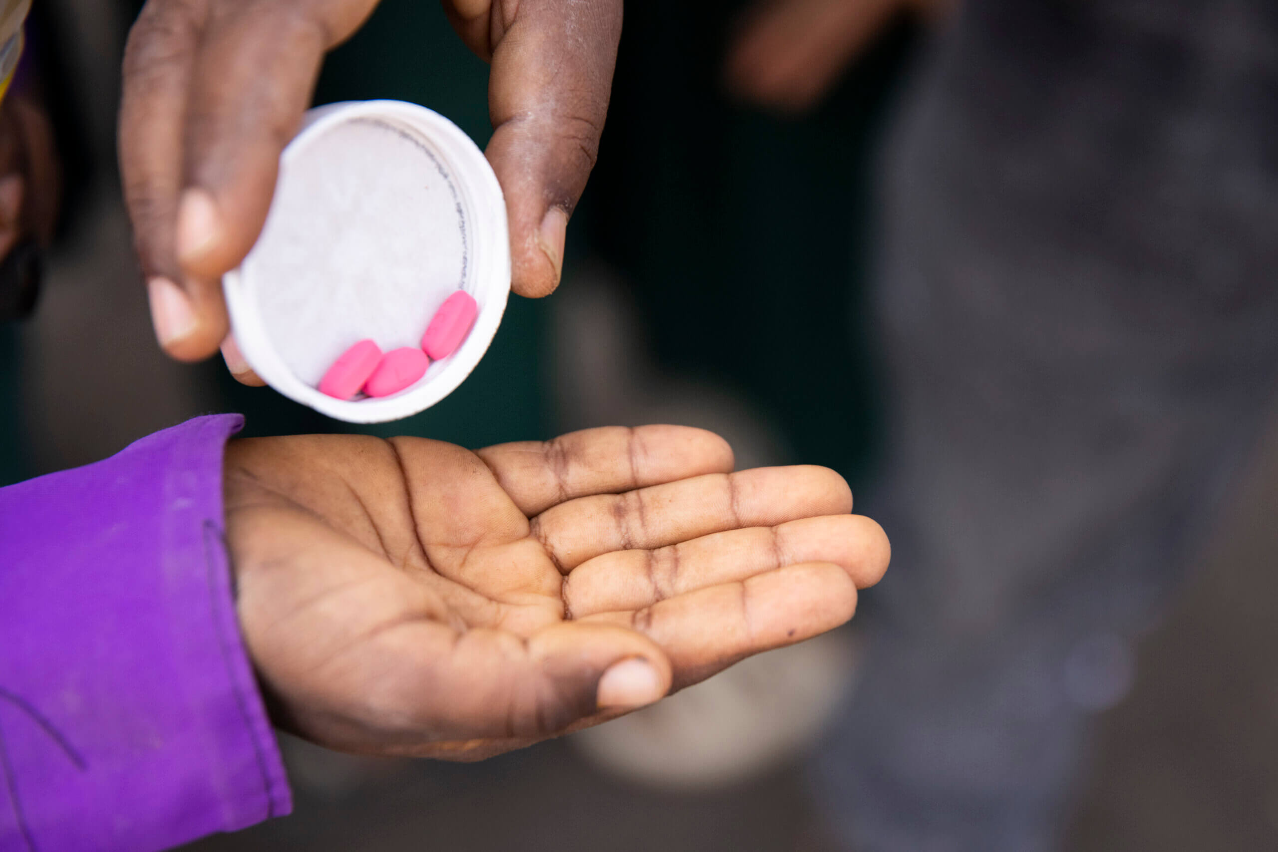 A child holds out their hand. They wear a purple top, you can see the sleeve. An adult tips some red tablets onto their palm, medication as part of a Child MDA (Mass Drug Distribution) in Mekoni in Tigray, Ethiopia. Investing in NTDs matters.