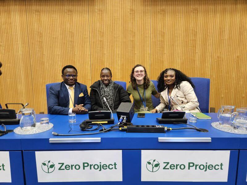 From left to right: moderator Ambrose Murangira, Senior Disability Inclusion Expert at Light for the World, with panellists Maryanne Emomeri, Innovator and Communication Specialist at United Disabled Persons of Kenya, Lydia Vlagsma, Policy Advisor at European Disability Forum, Olamide Mary Ewejobi, Disability Inclusion Facilitator at Joint National Association of Persons with Disabilities Nigeria. The four panellists sit at a large blue table, smiling. They have microphones in front of them, and signs that read "Zero Project".