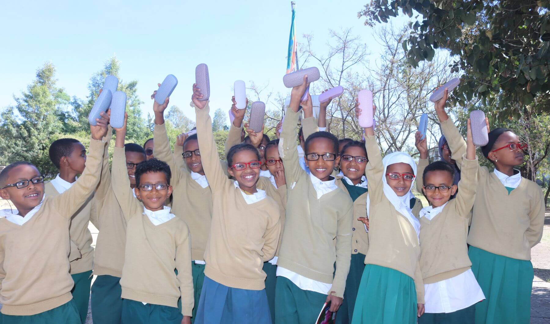 About 15 Ethiopian schoolchildren wearing school uniforms hold the cases for their glasses into the air. They are all wearing glasses and smiling.
