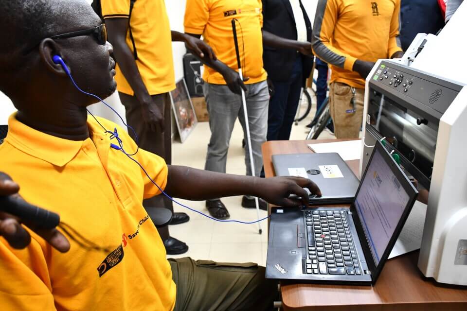 A Disability Inclusion Facilitator dressed in a yellow t-shirt sits at a computer to demoinstrate some of the assistive technologies available at the Inclusive Education Resource Centre as several others watch him.