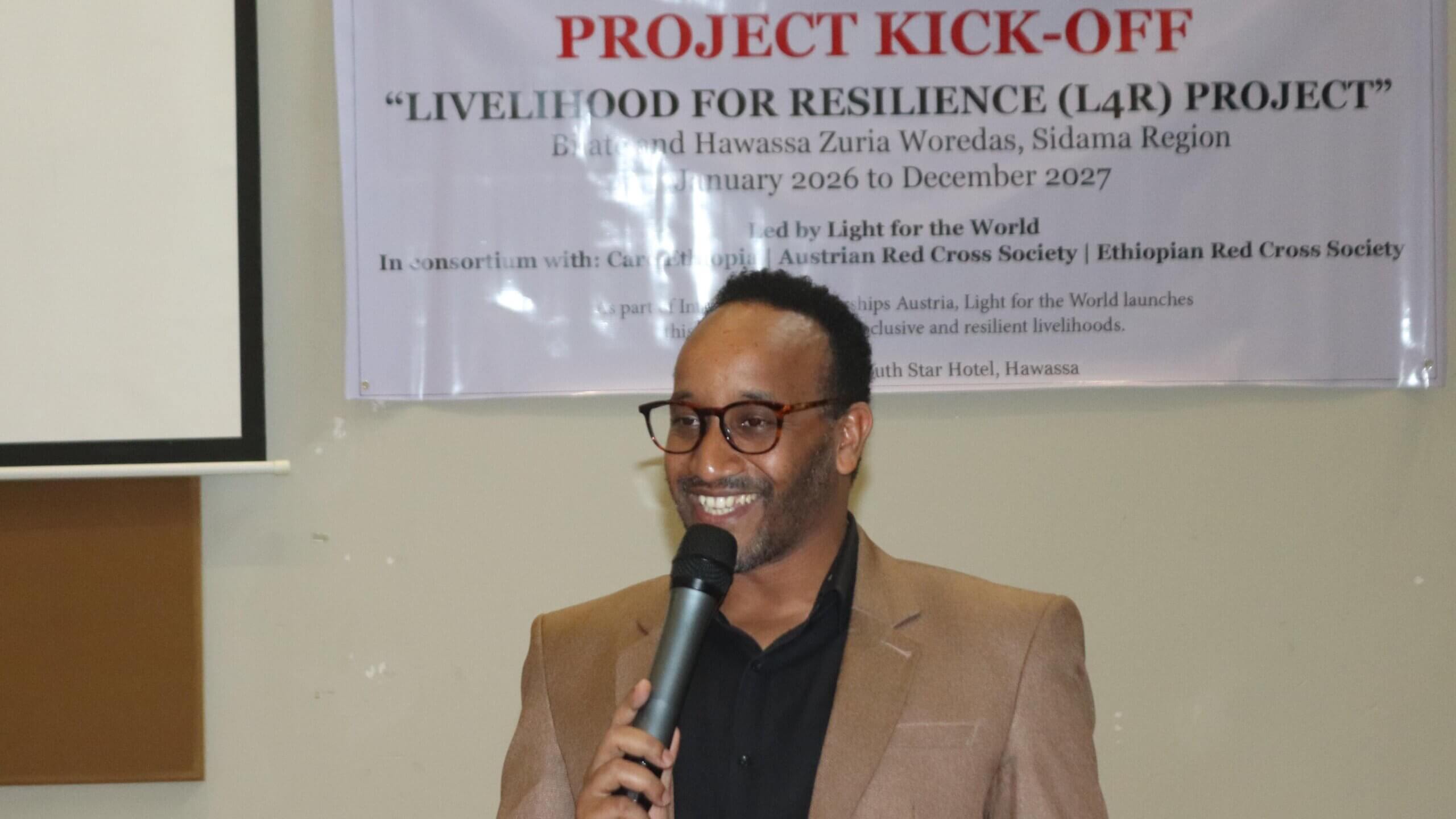 A man wearing glasses and a beige blazer speaks into a microphone at the Livelihoods for Resilience (L4R) project kick-off event. Behind him is a banner with details of the programme, which covers Bilate and Hawassa Zuria Woredas in the Sidama Region from January 2026 to December 2027.
