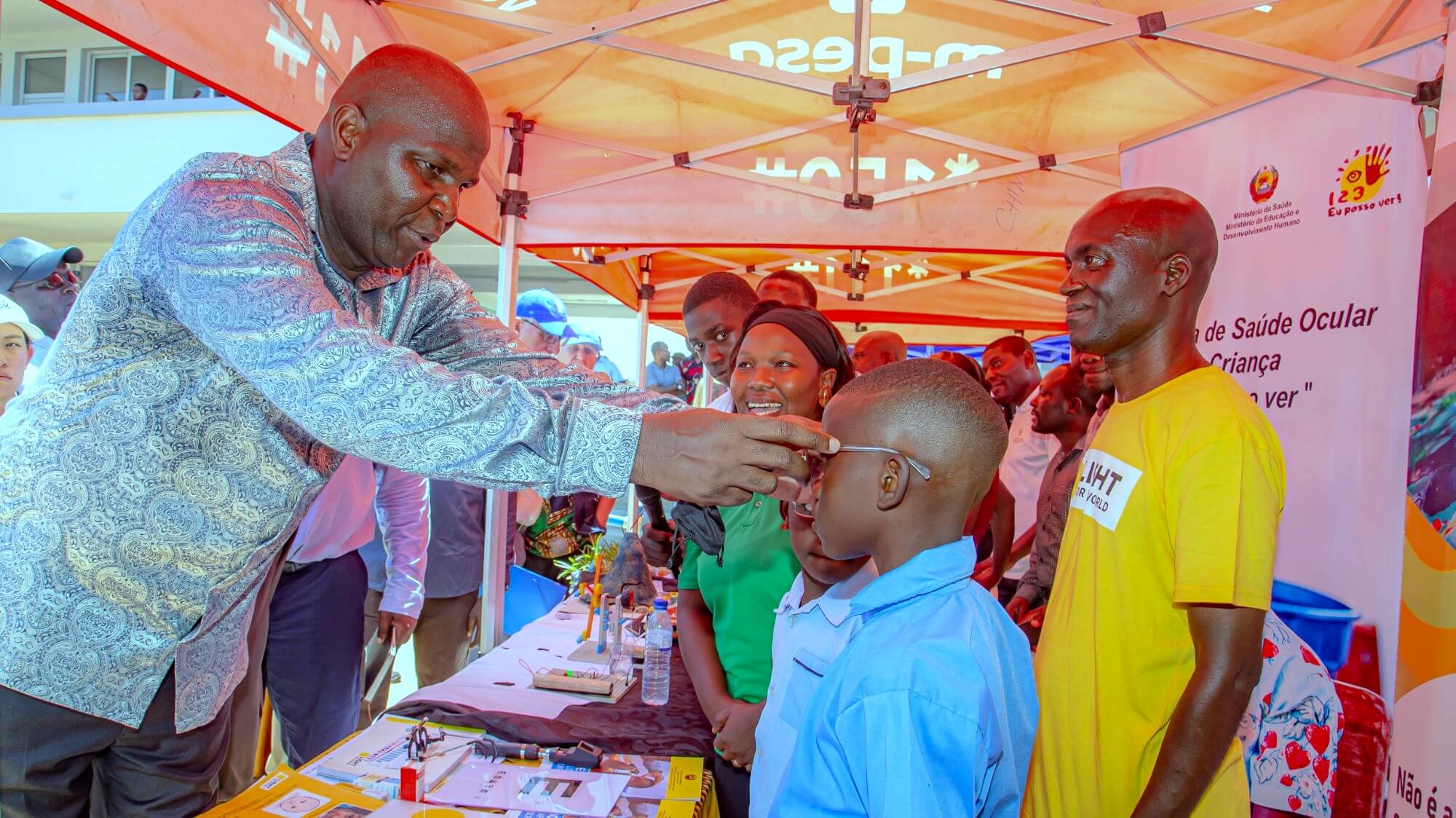 President Daniel Chapo places a pair of eyeglasses on a young boy in a blue school uniform at an outdoor exhibition stand. The stand features 1,2,3 Eu Posso Ver (I Can See) branding and a Light for the World banner. A woman smiles in the background and a man wearing a yellow Light for the World t-shirt stands beside the child.