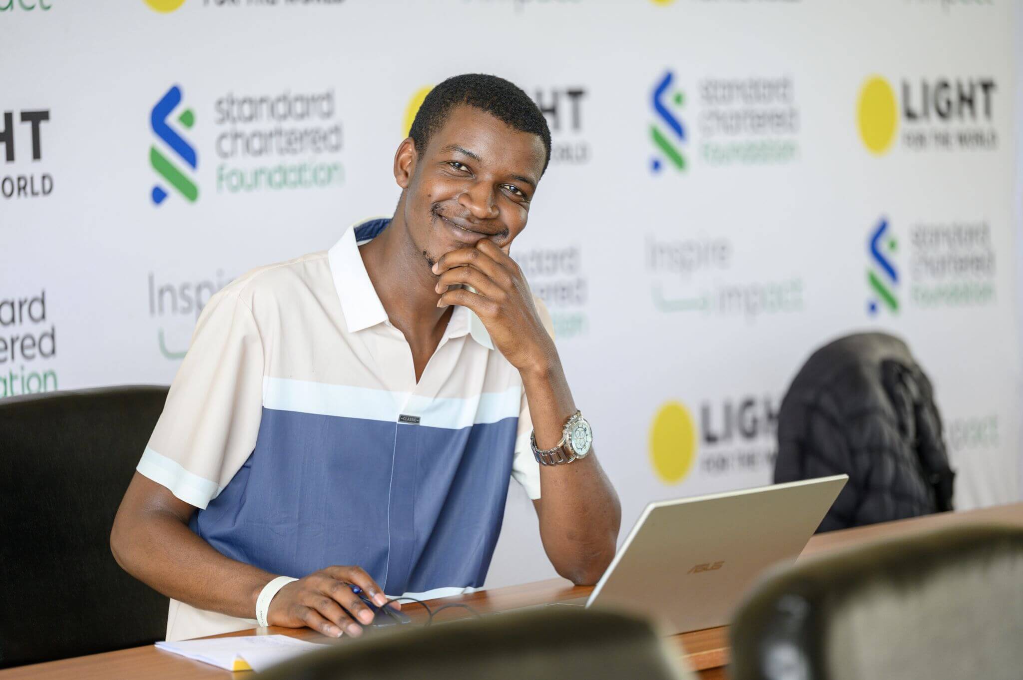 A young man sits at a desk with a laptop, resting his chin on his hand and smiling. Behind him is a branded backdrop featuring the logos of Light for the World, Standard Chartered Foundation and the words "Inspire Impact."