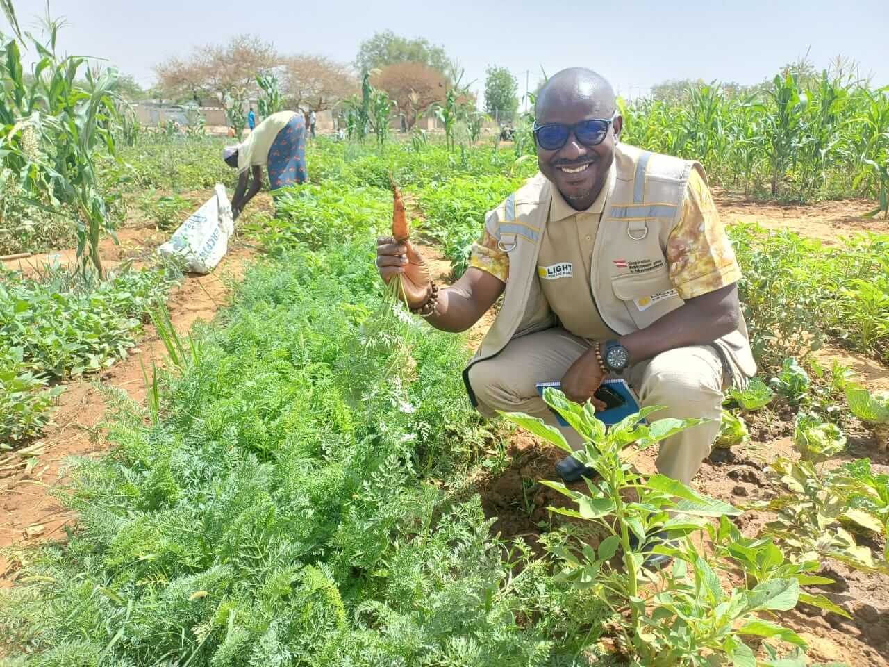 A smiling man wearing a Light for the World staff vest crouches in a vegetable garden and holds up a freshly harvested carrot. Green crops and another person working in the field are visible in the background.